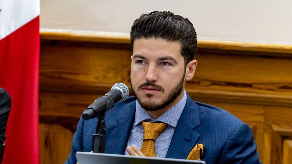 Man in a Blue Suit and Yellow Tie Speaks into a Microphone at a Formal Panel or Meeting Room. - Enbroncados