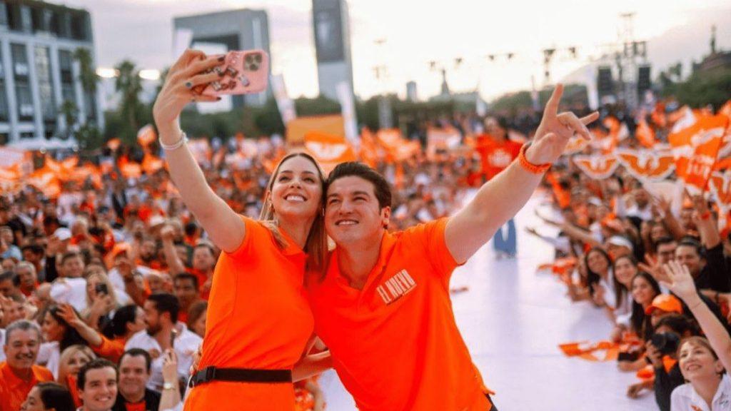 a Couple in Bright Orange Shirts Takes a Selfie with a Cheering Crowd Behind Them at an Outdoor Event. - Enbroncados