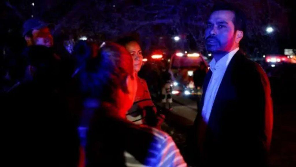 Nighttime Scene with a Man in a Suit Standing Among People As Police Cars Flash Red and Blue Lights in the Background. - Enbroncados