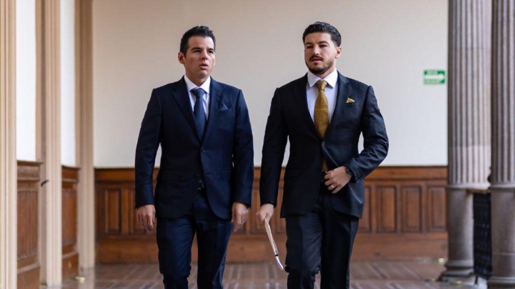 Two Men in Navy Suits Walk Down a Wood-paneled Hallway, One Holding Papers and Both Looking Ahead As They Talk. - Enbroncados