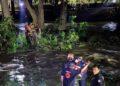 Emergency Responders in Gear Help a Person from a River at Night, with Others on the Riverbank Observing and a Tree-lined Backdrop. - Enbroncados