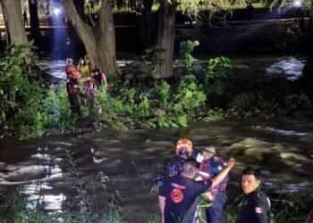Emergency Responders in Gear Help a Person from a River at Night, with Others on the Riverbank Observing and a Tree-lined Backdrop. - Enbroncados