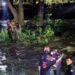 Emergency Responders in Gear Help a Person from a River at Night, with Others on the Riverbank Observing and a Tree-lined Backdrop. - Enbroncados