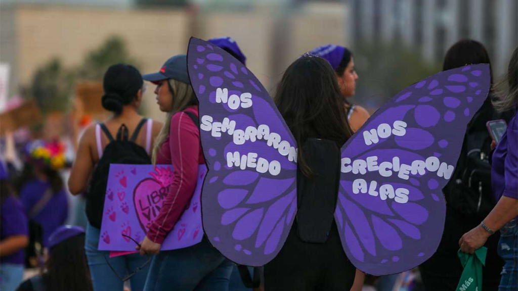 Person Wearing Purple Butterfly Wings with Spanish Text at a March; Crowd in Background. - Enbroncados
