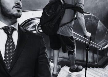 In grayscale, a man in a suit stands at the left while another person with a backpack and a crutch climbs down a stairwell behind him.