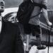 in Grayscale, a Man in a Suit Stands at the Left While Another Person with a Backpack and a Crutch Climbs Down a Stairwell Behind Him. - Enbroncados