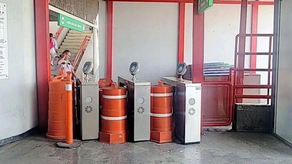 Samuel-garcia-no-muestra-la-realidad-del-metro 1 - Enbroncados Station Entrance with Three Metal Turnstiles and Orange Safety Barriers; a Person Stands Near the Left Gate Under Green Signage. - Enbroncados