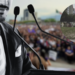 Public Speaker at a Podium Addressing a Crowded Outdoor Rally; a Purple Awareness Ribbon Overlays the Scene. - Enbroncados