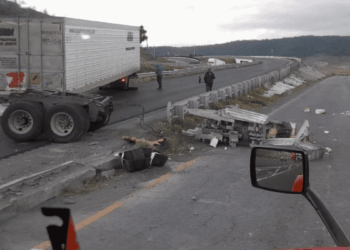 Semi-trailer Obstructing a Curved Highway with Scattered Debris and Damaged Guardrails; Two People Assess the Scene, with a Truck Side Mirror Visible in the Foreground. - Enbroncados